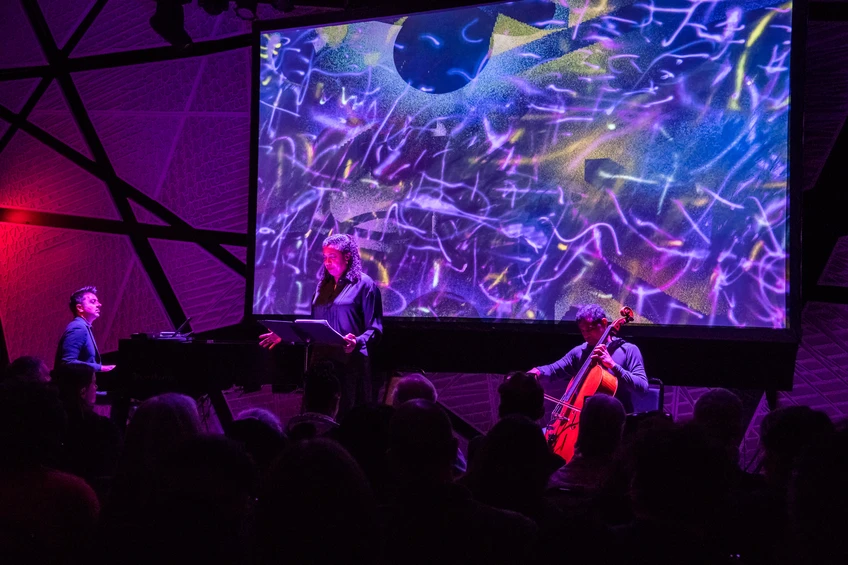 Three musicians performing on stage in front of a screen with artwork on it